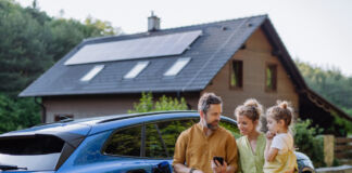 Eine Familie steht vor einem E-Auto. Dahinter liegt ihr Haus, auf dessen Dach Sonnenkollektoren angebracht sind. Der Vater zeigt der Mutter etwas auf seinem Smartphone. Sie lächelt und hält die kleine Tochter auf dem Arm. Währenddessen lädt das E-Auto.