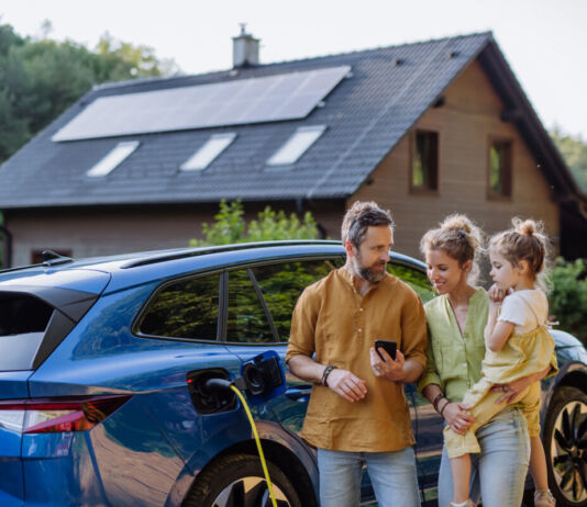 Eine Familie steht vor einem E-Auto. Dahinter liegt ihr Haus, auf dessen Dach Sonnenkollektoren angebracht sind. Der Vater zeigt der Mutter etwas auf seinem Smartphone. Sie lächelt und hält die kleine Tochter auf dem Arm. Währenddessen lädt das E-Auto.