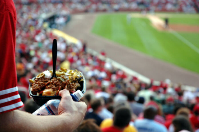 Ein Mann steht im Stadion und schaut sich ein Fußballspiel an. In der Hand hält er einen Behälter mit Nachos und Käsesoße als Snack während des Spiels. Im Hintergrund ist das Spielfeld zu sehen.