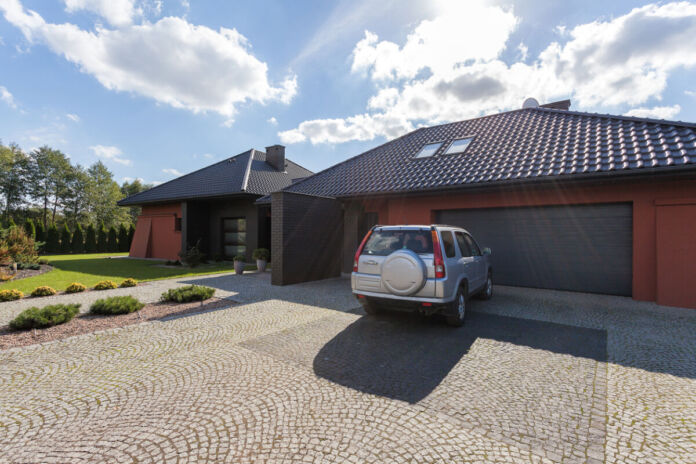 Ein großes silbernes Auto, ein Geländewagen, parkt vor einer privaten Garage. Daneben steht ein Einfamilienhaus. Davor ist ein sauberer kleiner Garten mit kurz geschnittener Wiese angelegt. Die Häuser sind Neubauten. Die Sonne scheint.