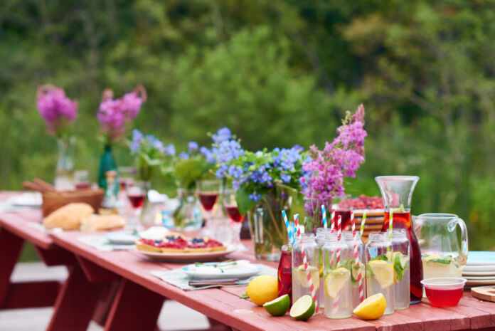 Ein Holztisch, der draußen in einem Garten steht, ist reichlich gedeckt mit Speisen und Getränken. In den vorderen Gläsern mit Trinkhalmen ist selbstgemachte Limonade. Der Tisch ist gedeckt und mit Blumen geschmückt. Ein beliebtes Getränk sollte man jedoch niemals zu lange offen stehen lassen.