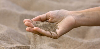 Der Sand an einem Strand fließt durch die Finger einer Frauenhand. Der Sand ist trocken, fein und frei von Steinchen und anderen Dingen, die man am Meer findet.