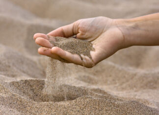 Der Sand an einem Strand fließt durch die Finger einer Frauenhand. Der Sand ist trocken, fein und frei von Steinchen und anderen Dingen, die man am Meer findet.
