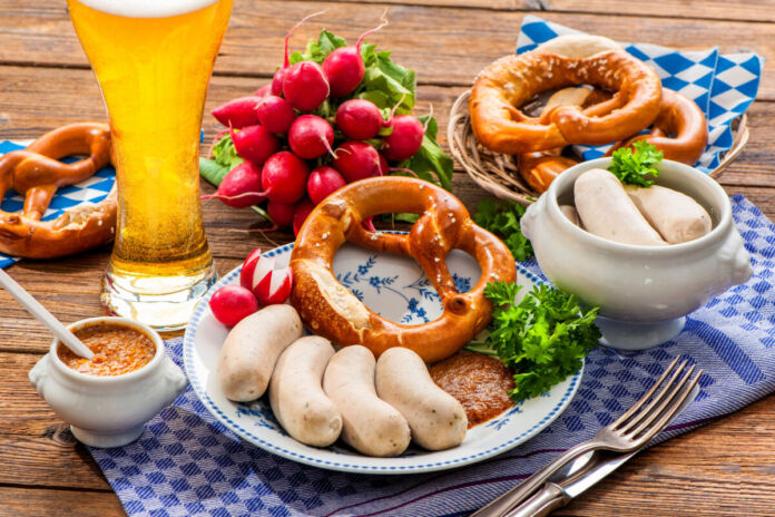Auf einem Tisch mit einer karierten Tischdecke steht Essen und Trinken. Dbei ist ein Bier, Brezel, mehrere Weißwürstchen, Senf und Radieschen. Das Essen wird auf einem Teller serviert.