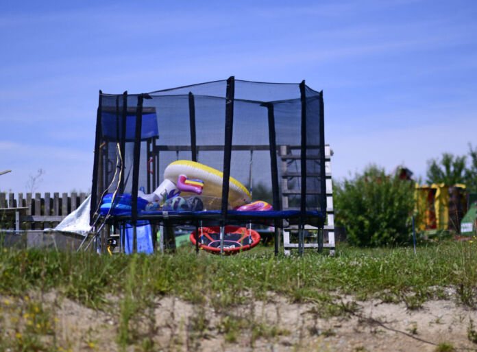 In einem großzügig umzäunten großen Garten steht ein großes Trampolin, das mit einem Netz umspannt ist. In diesem befinden sich viele Spielzeuge von Kindern. Im Hintergrund ist eine Nestschaukel angebracht.