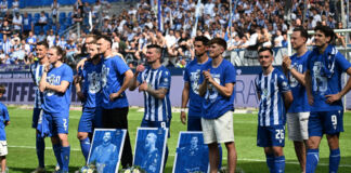 Die Spieler des Karlsruher SC stehen jubelnd und voller Vorfreude auf das Spiel nebeneinander im Wildparkstadion. Vor ihnen sind blaue Bilder von anderen Spielern, denen sie applaudieren. Dahinter stehen die KSC-Fans.