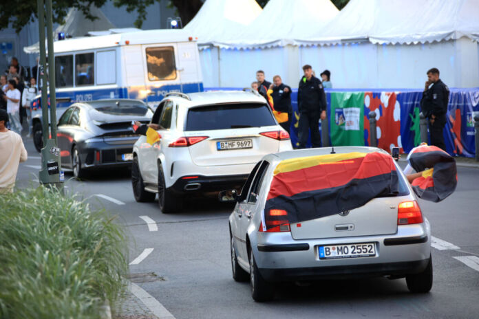 Verschiedene Autos mit Deutschlandflaggen als Dekoration fahren an Zelten vorbei. Neben den Zelten stehen Mitarbeiter vom Sicherheitsdienst, welche sich die fahrenden Fahrzeuge anschauen.