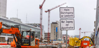 Eine große Baustelle ist abgesperrt. Im Vordergrund steht deshalb ein Baustellenschild mit der Aufschrift: Kein Durchgang. Passanten und Fußgänger dürfen diese Baustelle nicht betreten. Im Einsatz sind verschiedene Baukräne und Baugeräte.