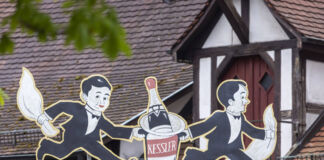 Auf einem Dach steht das Logo eines Traditionsunternehmens. Auf dem Logo der Marke sind zwei Männer zu sehen die eine Flasche Sekt tragen.