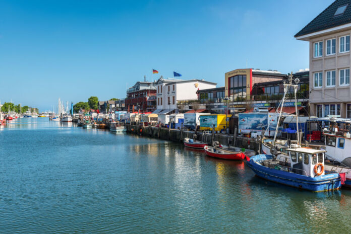 In einem Hafen am Wasser sind viele, bunte Boote angelegt. Daneben sind Häuser und eine Promenade zu sehen. Das Wasser ist blau.