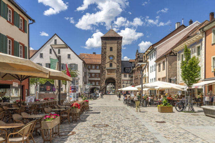 Eine gepflasterte Straße in der Fußgängerzone einer Stadt im Schwarzwald. Man sieht ein Café mit Stühlen und Tischen draußen. Am Ende steht ein Turm mit einem Tor, durch das die Menschen gehen können.