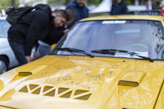 Das Close-up von einem gelben Kult-Auto, vermutlich ein Opel Manta. Ein Mann, ganz in schwarz gekleidet, begutachtet den Oldtimer, vielleicht auf einer Automobil-Messe.