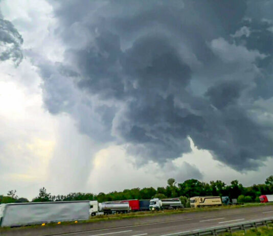 Am Himmel sind dunkle Wolken zu sehen. Es scheint, dass ein großer Sturm aufzieht. Im Hintergrund kann man vereinzelt sehen, wie es bereits regnet. Auf der Straße davor stehen viele LKWs in verschiedenen Farben.