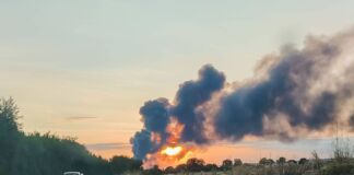 Bei einem Großbrand steigen dicke grau-schwarze Rauchwolken auf. Im Vordergrund ein Auto auf einer Straße. Der Brand ereignet sich in der Abenddämmerung, die Sonne geht gerade unter.