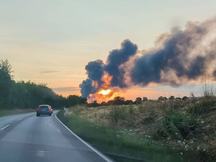 Bei einem Großbrand steigen dicke grau-schwarze Rauchwolken auf. Im Vordergrund ein Auto auf einer Straße. Der Brand ereignet sich in der Abenddämmerung, die Sonne geht gerade unter.