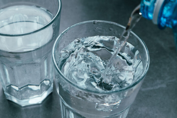 Auf einem Tisch stehen zwei Gläser voll mit Wasser. Jemand gießt in das vordere Glas noch etwas Wasser aus einer Flasche ein.