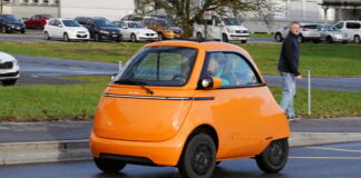 Ein Mini-E-Auto ist auf den Straßen unterwegs. Es ist der Microlino. Dieses Auto hat wenig Platz und keine Rücksitze. Das orange Elektroauto fällt sofort auf.