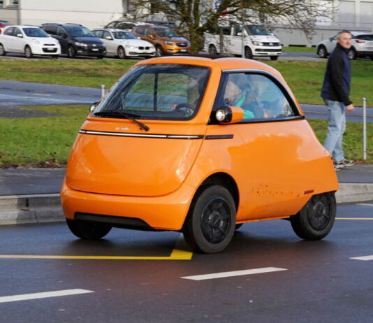 Ein Mini-E-Auto ist auf den Straßen unterwegs. Es ist der Microlino. Dieses Auto hat wenig Platz und keine Rücksitze. Das orange Elektroauto fällt sofort auf.