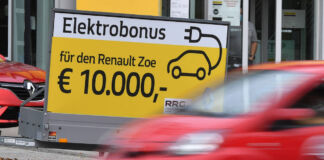 Ein rotes Auto fährt unscharf im Vordergrund. Dahinter ist ein großes gelbes Hinweisschild auf einem Anhänger. Es wirbt für einen Elektrobonus von 10.000 Euro für den Renault Zoe hin.