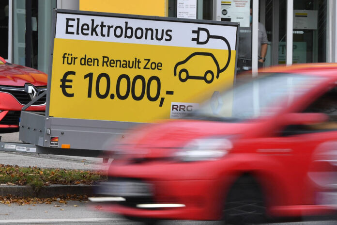 Ein rotes Auto fährt unscharf im Vordergrund. Dahinter ist ein großes gelbes Hinweisschild auf einem Anhänger. Es wirbt für einen Elektrobonus von 10.000 Euro für den Renault Zoe hin.