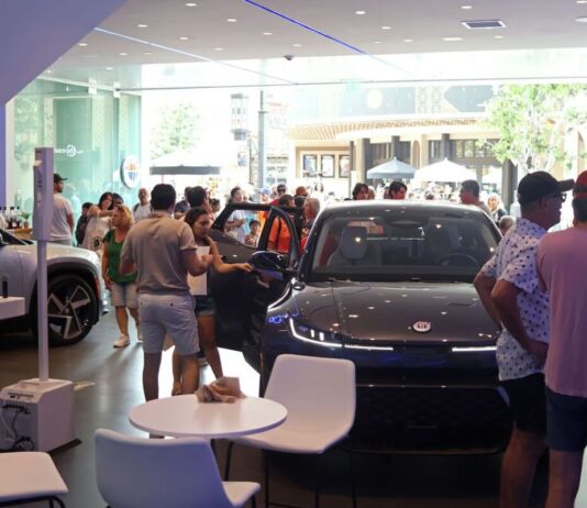 Mehrere Menschen feiern die Eröffnung eines neuen E-Autos in einem Showroom. In dem großen modernen Raum sieht man zwei luxuriöse Stromer sowie circa 20 Besucher, die sommerlich gekleidet sind. Hinter dem Schaufenster scheint die Sonne.