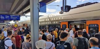 Zahlreiche Menschen stehen dicht aneinandergereiht auf einem überfüllten Zuggleis. Rechts im Hintergrund drängen sich viele Fahrgäste in einen Zug der Deutschen Bahn.