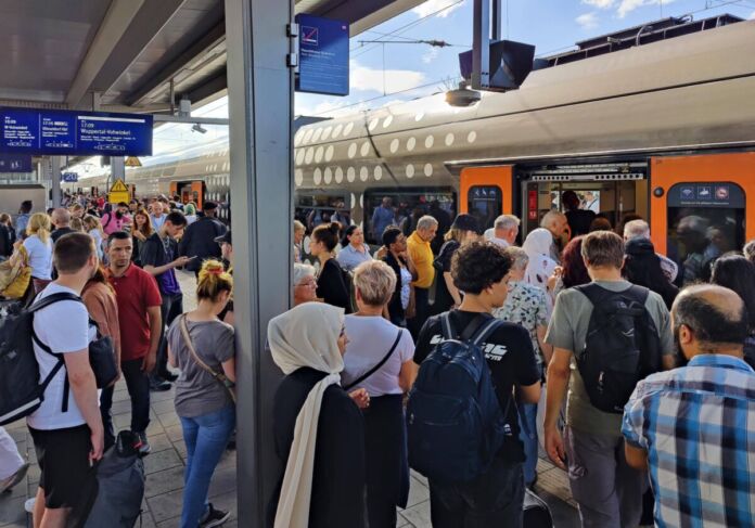 Zahlreiche Menschen stehen dicht aneinandergereiht auf einem überfüllten Zuggleis. Rechts im Hintergrund drängen sich viele Fahrgäste in einen Zug der Deutschen Bahn.