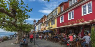 Neben dem Wasser sind verschiedene, bunte Häuser zu sehen. Darin sind Cafés und Restaurants eingerichtet. Draußen stehen viele Tische und Stühle, an welchen zum Teil Personen sitzen.