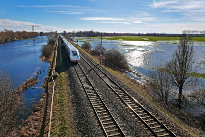 Ein ICE der Deutschen Bahn fährt durch eine überflutete Landschaft. Es scheint ein Hochwasser gegeben zu haben. Es ist sonnig und die Wiesen rechts und links von der Zugstrecke sind voller Wasser.