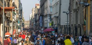 Auf einer Hauptstraße in einer Großstadt spazieren viele Menschen in unterschiedlicher Kleidung. Die Einkaufsstraße ist von Menschen angefüllt. Die Sonne scheint.