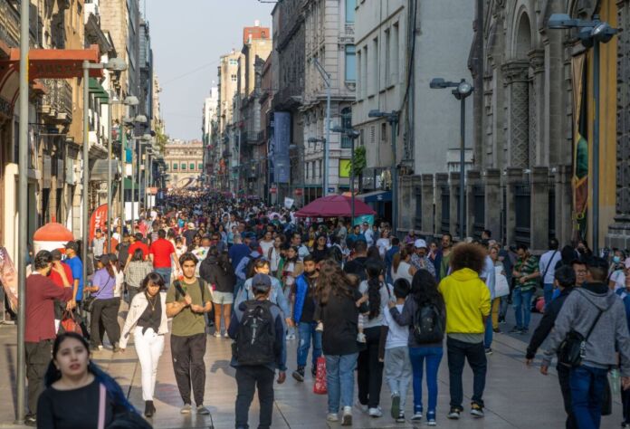 Auf einer Hauptstraße in einer Großstadt spazieren viele Menschen in unterschiedlicher Kleidung. Die Einkaufsstraße ist von Menschen angefüllt. Die Sonne scheint.