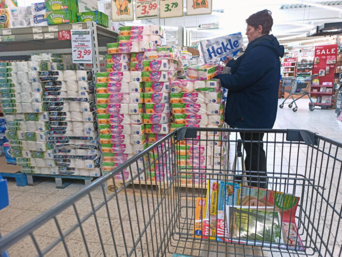 Eine Frau steht in einem Supermarkt oder Discounter vor einem riesigen Stapel voll Toilettenpapier. Sie hält zwei verschiedene Marken in der Hand und scheint offensichtlich zu vergleichen. Im Vordergrund erhält man einen Einblick in einen Einkaufswagen.