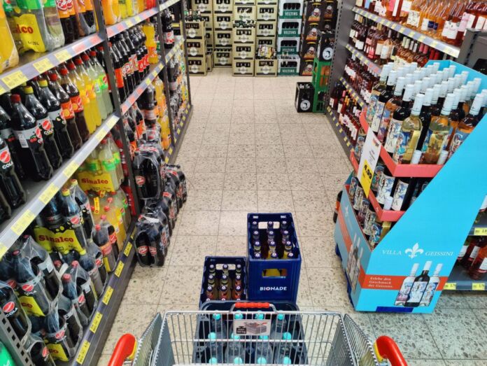 Ein Einkaufswagen wird durch die Getränkeabteilung in einem Supermarkt geschoben. In den Regalen stehen verschiedene Flaschen und auch Kisten mit Bier.