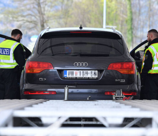 Ein Auto an der Grenzkontrolle. Man sieht den Pkw von hinten, Fahrer- und Beifahrertür stehen offen, davor jeweils ein Grenz-Polizist in gelber Warnweste. Offensichtlich überprüfen sie den Reisepass des Fahrzeugführers.