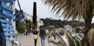Menschen spazieren neben Palmen an der Strandpromenade entlang. Große Hotelanlagen sind von Baustellenabsperrungen eingezäunt. Die Touristen sind bunt angezogen und die Palmen am Strand sind gelb von der Sonne.