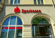 Eine Filiale der Sparkasse von vorne. Neben dem großen Schaufenster der Bank befindet sich der Eingang mit den elektrischen Türen. Im Inneren stehen die Geldautomaten und die Mitarbeiter warten auf die Kunden.