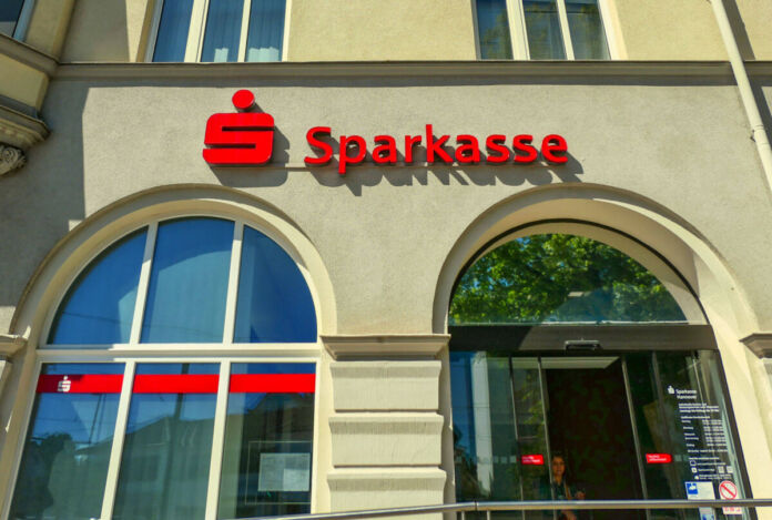 Eine Filiale der Sparkasse von vorne. Neben dem großen Schaufenster der Bank befindet sich der Eingang mit den elektrischen Türen. Im Inneren stehen die Geldautomaten und die Mitarbeiter warten auf die Kunden.