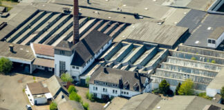 Eine große Produktionsstätte wird aus der Luft fotografiert. Das Werk liegt inmitten eines Industriegebiets. Die Dächer der Hallen sind grau.