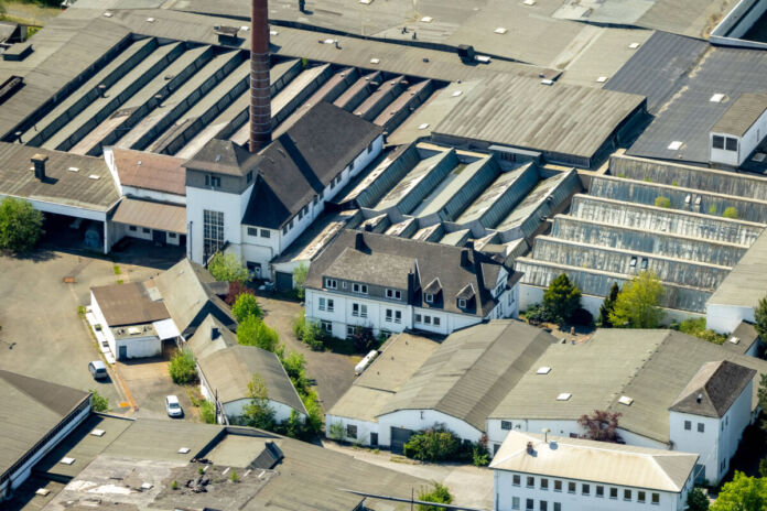 Eine große Produktionsstätte wird aus der Luft fotografiert. Das Werk liegt inmitten eines Industriegebiets. Die Dächer der Hallen sind grau. Ein Schornstein ist zu sehen und die gesamte Anlage scheint auf dem Land zu sein.