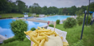 Eine Person hält eine Portion Pommes frites mit Mayonnaise in der Hand. Die Pommes hat er oder sie zuvor im Bistro des Schwimmbades gekauft. Im Hintergrund sind die Schwimmbecken zu sehen.