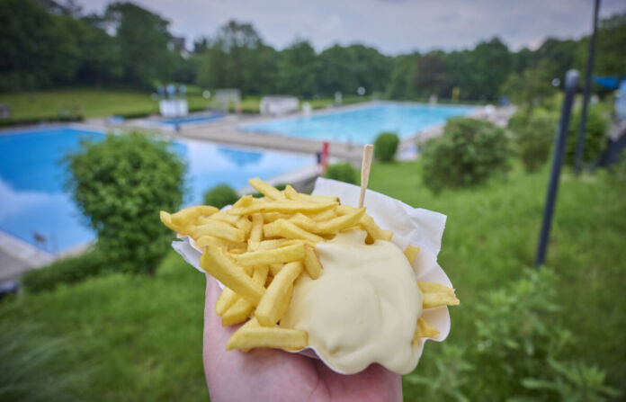 Eine Person hält eine Portion Pommes frites mit Mayonnaise in der Hand. Die Pommes hat er oder sie zuvor im Bistro des Schwimmbades gekauft. Im Hintergrund sind die Schwimmbecken zu sehen.