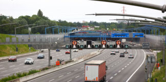 Eine mehrspurige, graue Autobahn mit verschiedenen Fahrzeugen. Auf der Straße fahren Autos und LKW. Oben sind leuchtende Straßenschilder zu sehen.