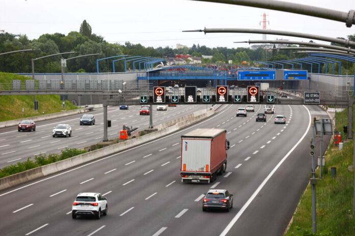 Eine mehrspurige, graue Autobahn mit verschiedenen Fahrzeugen. Auf der Straße fahren Autos und LKW. Oben sind leuchtende Straßenschilder zu sehen.