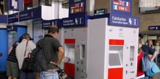 Personen stehen vor einem Fahrkartenautomat an einem Bahnhof. Ein Mann klickt auf die Schaltfläche des Automaten um sich ein Zugticket zu kaufen.