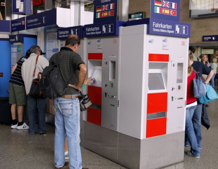 Personen stehen vor einem Fahrkartenautomat an einem Bahnhof. Ein Mann klickt auf die Schaltfläche des Automaten um sich ein Zugticket zu kaufen.