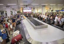 Nach ihrer Ankunft am Flughafen wartet eine große Menschenmenge am Gepäckband auf ihre Taschen und Koffer. Im Hintergrund hängt ein Bildschirm für Informationen für die reisenden Passagiere.