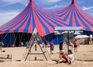 Im Hintergrund steht ein großes rot-blaues Zirkuszelt. Davor tummeln sich Kinder. Vermutlich finden in dem Zelt mehrere Veranstaltungen oder eine Großveranstaltung statt.