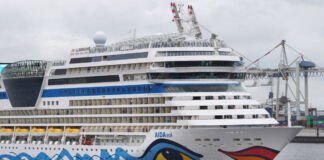 Ein gigantisches Kreuzfahrtschiff der Reederei und des Tourismus-Unternehmens Aida steht nah an einem Hafen. Grauer Himmel und graues Wasser sind zu sehen, ebenso wie Kräne des Hafens im Hintergrund.