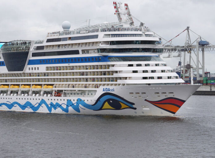 Ein gigantisches Kreuzfahrtschiff der Reederei und des Tourismus-Unternehmens Aida steht nah an einem Hafen. Grauer Himmel und graues Wasser sind zu sehen, ebenso wie Kräne des Hafens im Hintergrund.