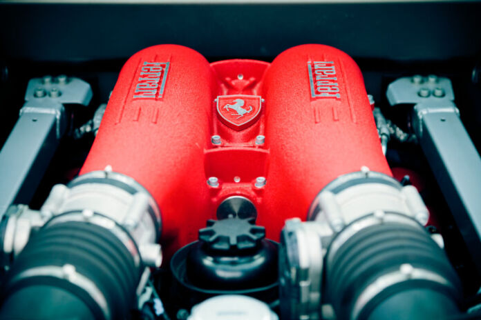 Ein leuchtend roter Motor ist in den Innenraum eines Autos eingepflegt. In der Mitte des Motors ist ein hochwertiges Schild und Logo des Herstellers Ferrari zu sehen, ein weißes aufbäumendes Pferd. Links und rechts prangen Schriftzüge des Herstellers den schönen Motor.